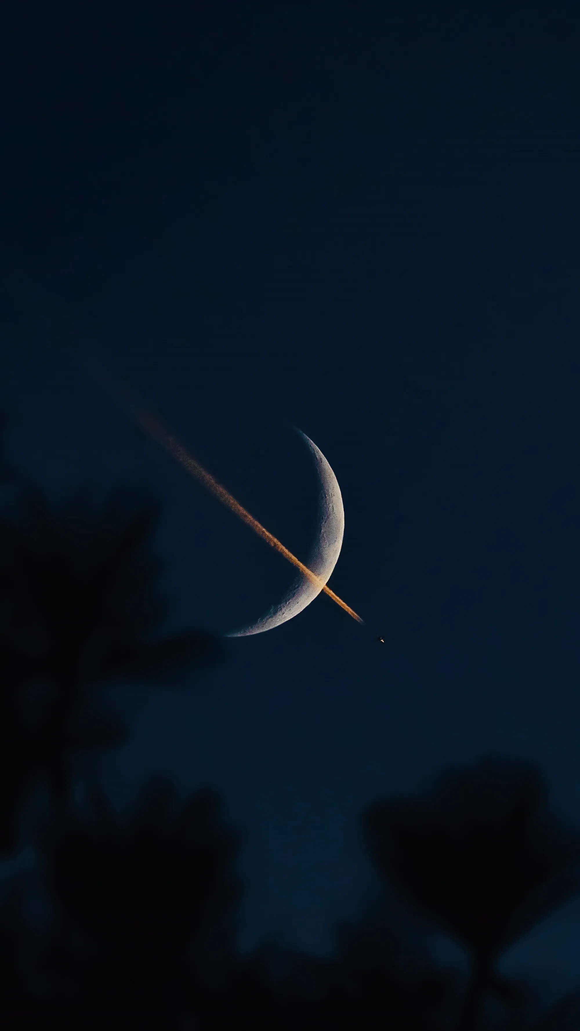 A sharp crescent moon bisected by a high-altitude jet contrail catching the last golden light of twilight.